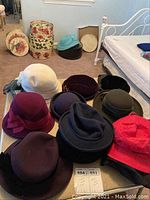 Wide shot showing 13 vintage women's hats in different colors and styles displayed on a surface and floor, with a grouping of six decorative vintage hat boxes in background.