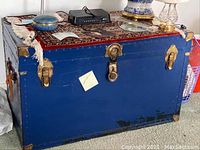 Front view of a blue wooden chest with brass metal trim and lock, showing scratches and a rug on top.