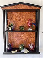 Full view of wooden wall-mounted display shelf holding six glass paperweights including fish, apples, rooster, and birds.