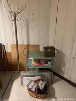 Photo showing metal clothes tree rack, metal rolling cart with rust, wicker basket with items, and miscellaneous items including packaged smoke alarms and flashlights.