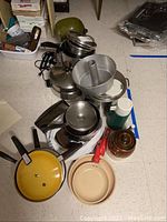 Wide view of group of cookware including pots, pans, cake pan, Thermos, and ceramic canister on floor