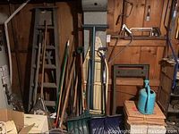Photo showing wooden ladder, ironing board, metal dolly, and various yard tools like shovels and rakes leaning against the wooden wall.