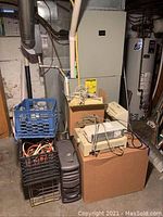 Pile of electronics in basement including Dell computer tower, crosscut shredder, printers on top of brown cardboard box, speakers, and milk crates with cords.