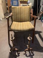 Front view of vintage arm chair with stained fabric upholstery and wooden frame. Pair of decorative metal fireplace irons placed in front on floor.