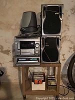 Overall view of AIWA stereo system setup including stereo unit, speakers, and metal cart with CDs and cassettes below