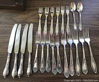 Full view of the 26 flatware pieces arranged by type on a wooden surface, showing knives, forks, teaspoons with detailed handle designs