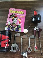 Photo showing the retro cocktail book, golf club-shaped cocktail tools including strainer, bottle opener, muddler and two golf tee toothpicks with holder, and a red and black leather pouch.