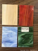 Four stained glass pieces of different colors and patterns displayed on a wooden table. Colors include creamy beige, deep red-orange, light blue with white swirls, and dark green with white swirls. One piece labeled Spectrum Glass Co.