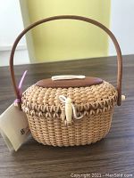 Front and side angle showing overall shape, handle, and attached tag visible. Basket oval with wooden top and bottom, wooden handle, and woven body.