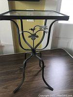 Full view of the black wrought iron garden table with square glass top on a wooden surface.