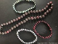 View of four freshwater pearl pieces on black background, including one long deep maroon pearl necklace and three bracelets in pale green, maroon, and lilac colors.