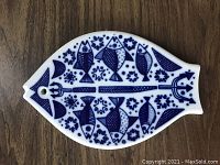 Front view of leaf-shaped ceramic trivet with blue fish and floral designs on white background.