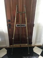 Front view of tall decorative brass easel standing indoor against wooden door, showing overall frame and height.