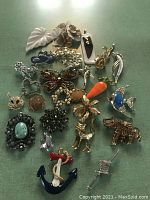 Top view of 25 assorted colorful vintage pins with various animal, insect, fruit, and flower motifs on a green background.