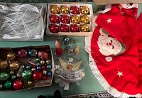 Photo showing a large box of clear plastic icicles, two boxes of vintage glass bulbs in gold, red, and other colors, a red felt Santa tree skirt with white fringe, assorted figurine ornaments including angels, and wooden decorations.