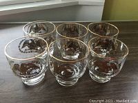 Six clear short glasses with gold rim and American eagle motif arranged on countertop.