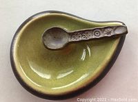 Top and angled view of an avocado green tear-shaped pottery serving dish with a small ceramic spoon inside. The dish has dark brown edges and a smooth glazed finish.