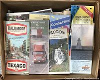A box filled with folded vintage highway maps and tourist pamphlets from various U.S. states and regions including Baltimore, Pennsylvania, Connecticut, Oregon, Maine, and Nova Scotia.