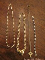 Four pieces of gold toned costume jewelry laid out on a wooden surface: two necklaces on left (beaded and chunky heart necklace), delicate necklace with Celtic cross pendant, and a bracelet with alternating black and white stones on the right.