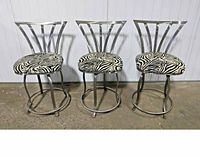 Front view of three brushed steel swivel bar stools with zebra print seats and curved vertical backrests against white paneled wall.