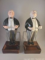 Front view of two vintage ceramic butler figurines standing on rectangular bases with card holder racks. Each figurine is dressed in a tuxedo and striped pants, smiling with open hands.