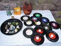 Photo showing a round black tray with Dog Patch cartoon characters, 12 assorted 45 RPM records, yellow advertising bank, wooden owl bank, and ceramic candle holder on cloth.