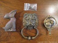 Overhead view of three brass figural door knockers: pelican on left, lion center with attached knocker ring and mounting screws above, kissing sweethearts on right with circular ring