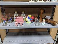 Wide photo of vintage kitchen and dining table items arranged on a metal shelving unit. Includes bird water tumblers, figural milkmaid salt and pepper shakers, wooden pepper mills, ceramic rooster spoon holder, mushroom salt and pepper shakers, napkin holder, and napkin ring holder.