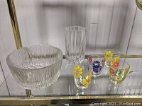 View of all items in lot: textured clear glass bowl, tall vase with vertical ridges, and 4 floral juice glasses arranged on glass shelf