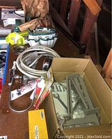 Lot of household hardware items including shower hose and head, toilet seat cover, power outlet, bulbs, and landline phone set components, displayed on table and in cardboard box.