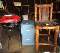 Photo showing a red portable grill on black metal frame with wheels, adjacent to a blue metal two-drawer filing cabinet, and a solid bamboo stool with woven seat and backrest.