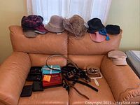 Wide shot of multiple hats positioned behind and along couch backrest with sunglasses, belts, small mirror, and beads arranged on seat cushion.