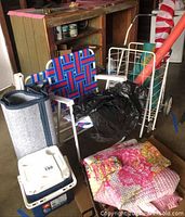 Photo showing picnic chairs with blue and red woven fabric, steel shopping cart with white frame, multicolored blankets stacked on cardboard, Igloo cooler, and a rolled orange yoga mat in background.