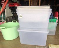 Seven clear plastic storage bins stacked, showing translucent material and measuring approx 22x16x13 or 23x16x13 inches. One green round tub visible to the left with handles.