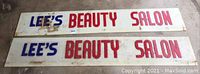Two rectangular acrylic signage plaques with blue and red lettering reading 'LEE'S BEAUTY SALON' laid flat on floor, showing dirt and stains needing cleaning.