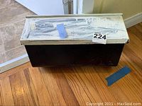 Front view of black metal mailbox with white lid closed, placed on wooden floor near doorway.