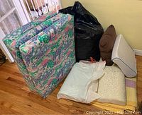 Photo showing the folded colorful mattress along with pillows, black and white plastic bags that likely hold a comforter and a quilt, and an off-white patterned cushion.