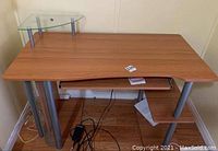 Front angled photo of the computer table showing the wood grain top, small glass elevated shelf on the left back, pull-out keyboard tray below, metal legs and vertical mesh column on left side.