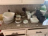 Kitchen counter with multiple mugs, bowls, plates including white ceramic bowls, small stainless steel bowl, white square plates and various mugs.