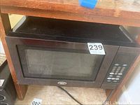 Oster microwave shown in its entirety, featuring stainless steel front, transparent door, and digital keypad on right side. Power cord visible below.