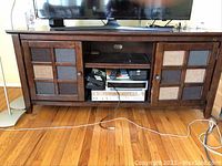 Front view of wooden entertainment console table with TV on top, showing paneled doors and open shelves with electronic items inside.