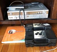Karaoke DVD player on wooden shelf with remote control on top, wired microphone draped over it, two black microphone cases, and an orange song book placed on the floor in front.