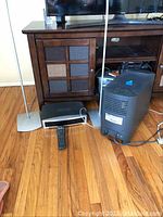 Photo showing all the system components on the floor under a wooden console, including the subwoofer, speakers, control unit, and remote.