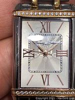 Close-up of watch face showing silver dial, rose gold Roman numerals, and crystal accents on bezel above and below dial.