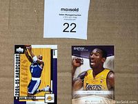 Both Kobe Bryant basketball cards placed on brown surface, one from Upper Deck 2004-05 Hardcourt, showing Kobe in blue Lakers jersey jumping for a shot, and the other 2021 Skybox Autograph card with Kobe celebrating in yellow Lakers jersey with printed signature.