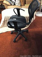 Side view of black office chair showing mesh backrest, seat, armrests, and caster wheels on brown carpet.