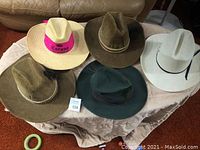 Overview of 5 cowboy/cowgirl hats in various colors and materials, placed on a beige cloth