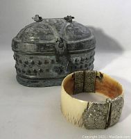 Photo showing the carved bracelet beside the antique metal box, both items are in focus with visible surface details.