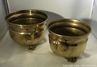 Two polished brass footed pots with handles, viewed from the side and angled to show handle details and rounded shape. Larger pot is on the left, smaller on the right.