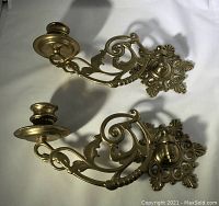 Pair of brass wall mount candle holders laid flat showing elaborate scrolling design and mounting plate details.